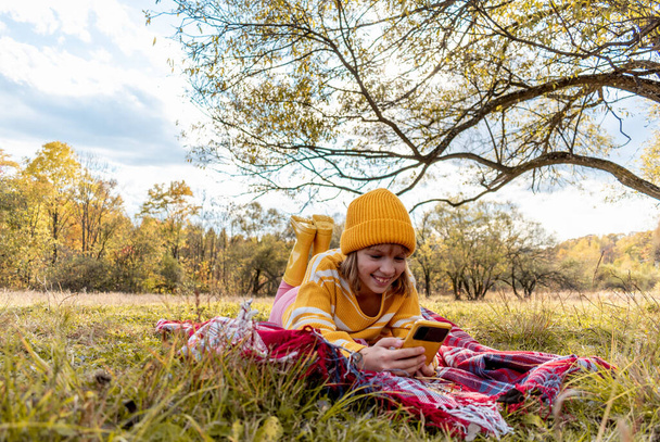 Happy girl takes video on the camera of his smartphone. Cozy slow lifestyle concept. Picnic in the meadow next to the tree. Warm up with hot drink during an autumn. - Фото, изображение
