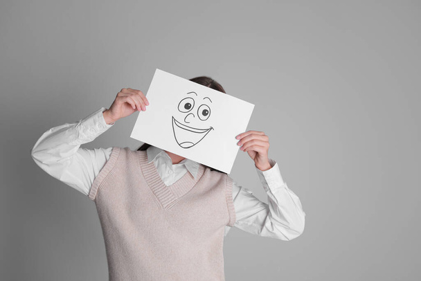 Woman hiding behind sheet of paper with happy face on grey background, space for text - Foto, imagen