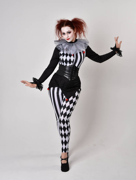 Portrait complet de fille aux cheveux roux portant un costume de bouffon de clown noir et blanc, personnage de cirque théâtral. pose debout isolée sur fond de studio. - Photo, image