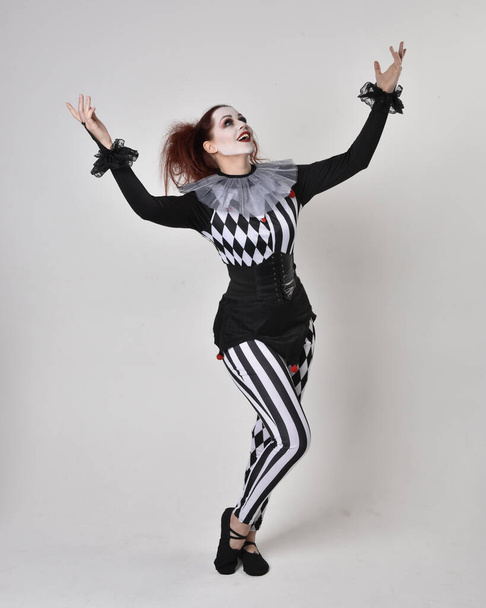 Volledig portret van een roodharig meisje in een zwart-wit clown nar kostuum, theatraal circusfiguur. Staande houding geïsoleerd op studio achtergrond. - Foto, afbeelding