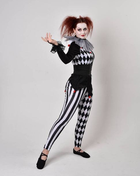 Portrait complet de fille aux cheveux roux portant un costume de bouffon de clown noir et blanc, personnage de cirque théâtral. pose debout isolée sur fond de studio. - Photo, image