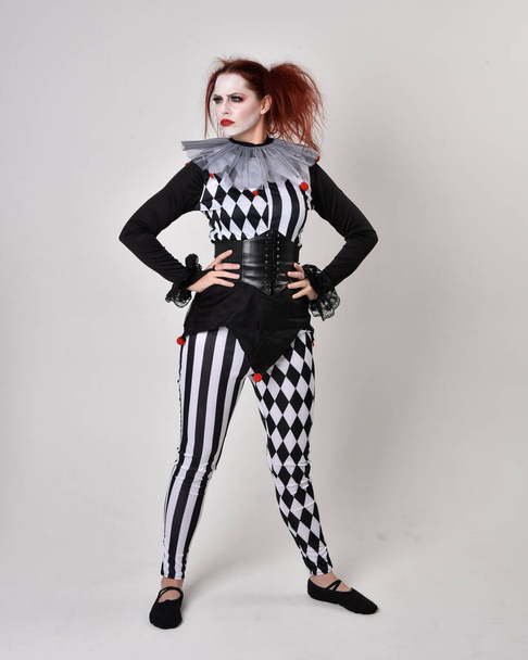 Volledig portret van een roodharig meisje in een zwart-wit clown nar kostuum, theatraal circusfiguur. Staande houding geïsoleerd op studio achtergrond. - Foto, afbeelding