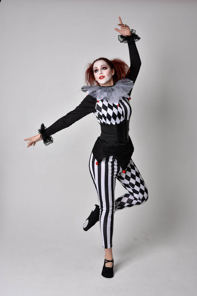 Retrato de cuerpo entero de una chica pelirroja vestida con un traje de bufón de payaso blanco y negro, personaje teatral de circo. pose de pie aislado en fondo de estudio. - Foto, imagen