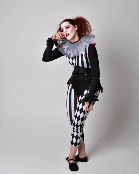 Full length  portrait of red haired  girl wearing a black and white clown jester costume, theatrical circus character.  Standing pose  isolated on  studio background. - Φωτογραφία, εικόνα