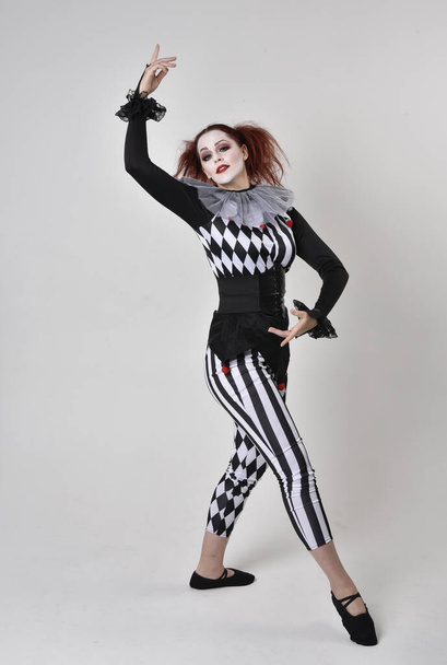 Ganztägiges Porträt eines rothaarigen Mädchens, das ein schwarz-weißes Clownskostüm trägt, eine theatralische Zirkusfigur. Stehende Pose isoliert auf Studiohintergrund. - Foto, Bild
