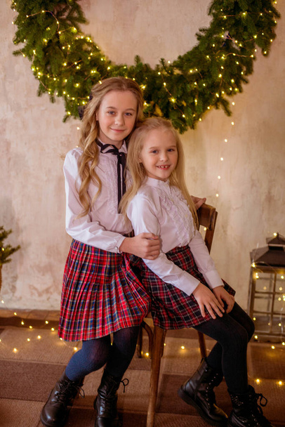 Lovely girls sisters with blonde curly hair with bans to the tols in classic school clothes  next to a large Christmas wreath of pine needles on the wall  in a classic interior. Christmas mood  - Foto, Bild
