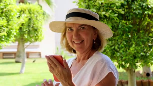 ξανθιά σενιόρα γυναίκα, 50-55 ετών, με καπέλο και λευκό φόρεμα, μιλά χρησιμοποιώντας ένα smartphone μέσω βιντεοκλήσης, πληκτρολογώντας ένα μήνυμα, αναζητώντας πληροφορίες στο διαδίκτυο στη βεράντα της ένα ηλιόλουστο πρωινό - Πλάνα, βίντεο