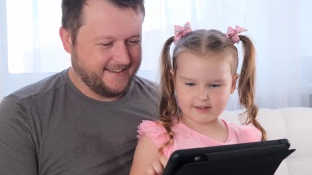 naurava pikkutyttö 3-vuotias ja hänen isänsä opiskelevat yhdessä tabletilla ja katsovat oppitunnin verkossa kotona. Isä auttaa tytärtään hänen opinnoissaan. Verkossa tapahtuvan etäopiskelun käsite. - Materiaali, video