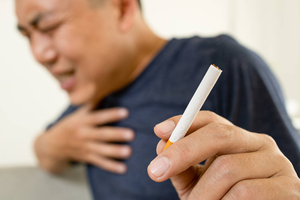 Asiatische männliche Raucher halten eine Zigarette, berühren seine Brustschmerzen mit der Hand, Atembeschwerden, Erstickung durch Rauchen, Mann umklammert seine Brust mit akuter Atemnot, Herzinfarkt, Herz- oder Lungenkrankheiten Konzept - Foto, Bild
