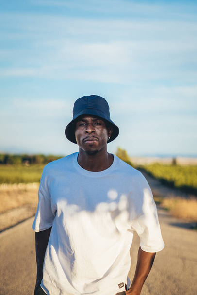 A portrait of a young black handsome man wearing a bucket hat walking along the highway at sunset - Фото, зображення