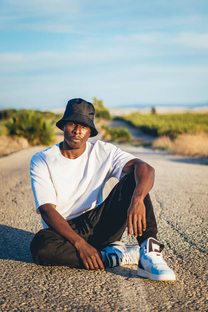 A young black handsome man wearing a bucket hat sitting on the ground in the middle of a road, sunset - Φωτογραφία, εικόνα