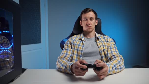 Молодой человек играет в компьютерную игру с геймпадом в полутемной комнате - Кадры, видео