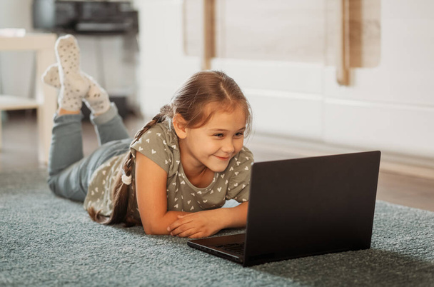小さな女の子がコンピュータ画面の前のカーペットの上に横たわっている。オンラインコミュニケーション - 写真・画像