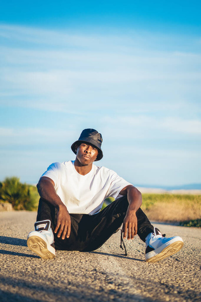 A young black handsome man wearing a bucket hat sitting on the ground in the middle of a road, sunset - Fotoğraf, Görsel