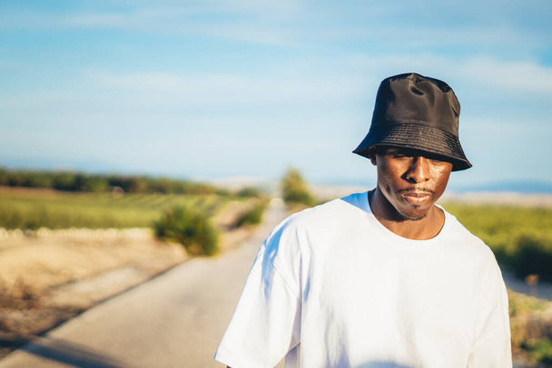 A portrait of a young black handsome man wearing a bucket hat walking along the highway at sunset - Foto, imagen