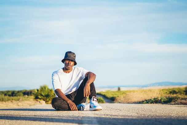 A young black handsome man wearing a bucket hat sitting on the ground in the middle of a road, sunset - Foto, Bild