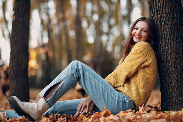 黄色の葉を持つ秋の公園の若い女性モデルカエデの葉。秋のファッション. - 写真・画像