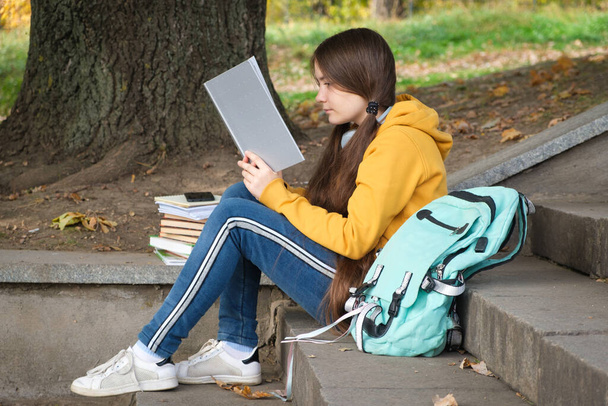 A girl of 13 years old sits on the stairs and reads a book in the park, goes to school exams. - Foto, imagen