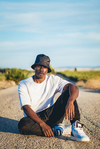A young black handsome man wearing a bucket hat sitting on the ground in the middle of a road, sunset - Photo, image