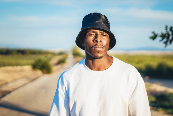 A portrait of a young black handsome man wearing a bucket hat walking along the highway at sunset - Foto, Bild