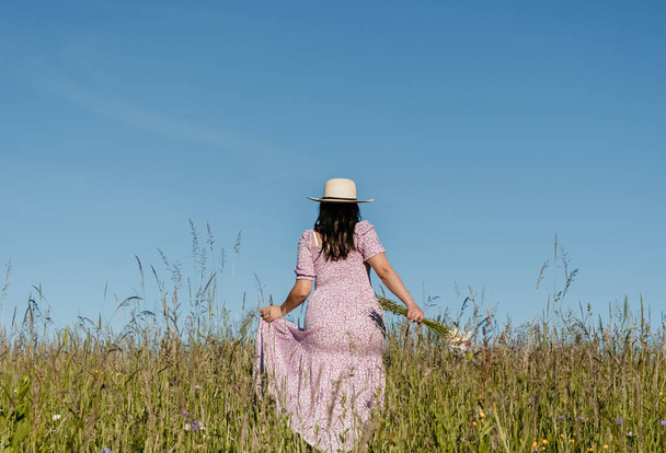 Lifestyle image of beautiful woman with long hair, walking in field or meadow, holding wildflowers. - Fotó, kép