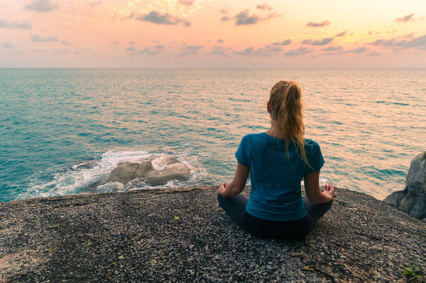 Young woman in lotus position meditates on rocks by the sea at sunrise. Colorful sky on the background. - 写真・画像