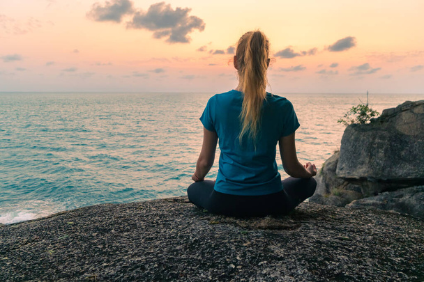 Jovem mulher em posição de lótus medita sobre rochas junto ao mar ao nascer do sol. Céu colorido no fundo. - Foto, Imagem