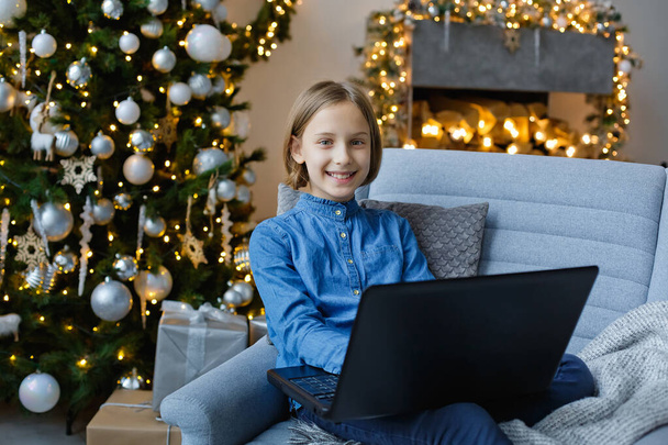 A cute girl is sitting on a sofa with a laptop in a decorated room for Christmas. Communication, meetings and training online. Merrily looks at the camera. - Photo, Image