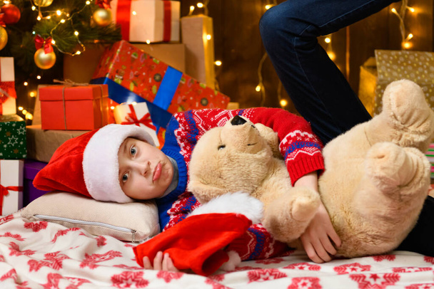 Kind Mädchen posiert im neuen Jahr oder Weihnachtsdekoration. Weihnachtslichter und Geschenke, mit Spielzeug geschmückter Weihnachtsbaum. Sie trägt einen roten Pullover und eine Nikolausmütze - Foto, Bild