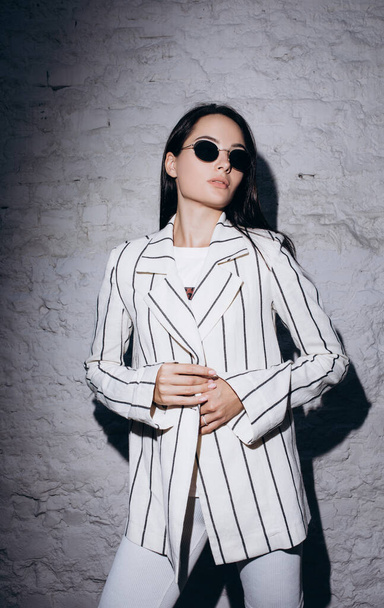 Young woman in sunglasses stands near brick wall dressed in striped white jacket and shorts. - Foto, Imagen