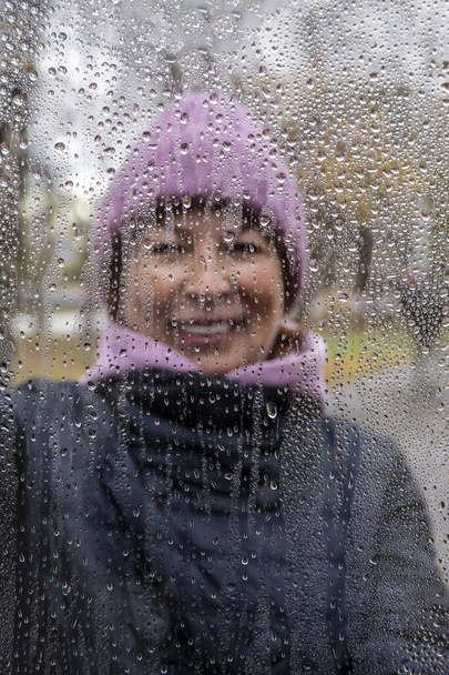 mujer en sombrero púrpura a través de vidrio con gotas de lluvia sobre fondo de otoño  - Foto, Imagen