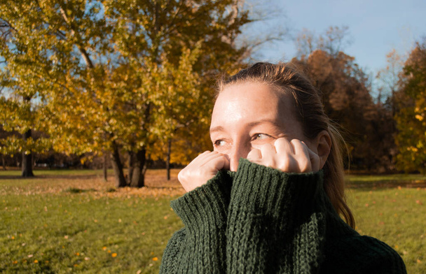Une femme est timide de la caméra, cache le visage dans le col d'un pull vert, fond de parc d'automne. - Photo, image