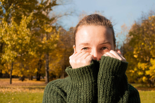 Woman looks at camera, hides her face in the collar of a green sweater, background of an autumn park. - Photo, Image
