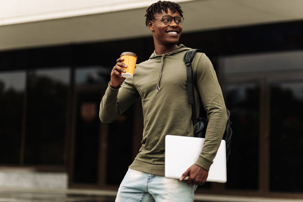 Stylish smiling African American man in glasses, drinking coffee in a cup and walking up the stairs in the city center with a laptop in his hands, stylish freelance people, with a laptop - Foto, immagini