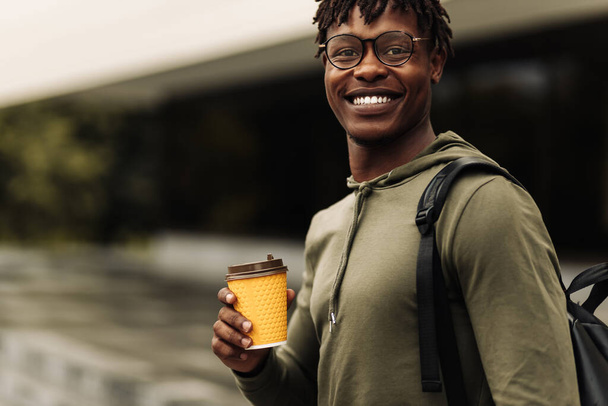 Coffee break for student, handsome African American male student, wearing glasses and with a backpack on his shoulder, drinking coffee on the street in the city - Photo, image