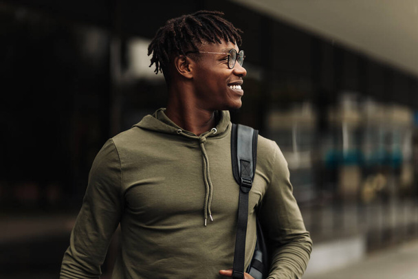 Happy handsome successful african student, wearing glasses and with a black backpack, standing on the steps and smiling, looks into the frame outdoors on the street near the university - Foto, immagini