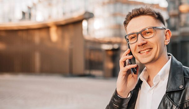 Handsome man with glasses with a smartphone on the street of a big city. Businessman talking on the phone on urban background - Photo, image