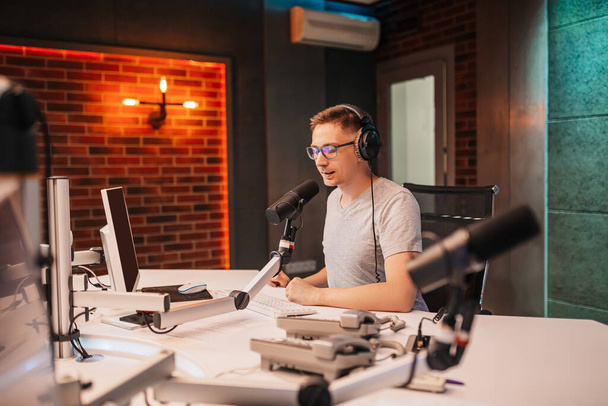 The DJ hosts the program and communicates with the audience on air at the radio station. The announcer reads the news. A male radio host speaks into a microphone and records a podcast - Foto, Imagem