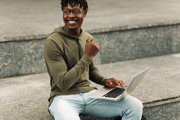 Happy Black African man, with glasses working on laptop outdoors, curly haired man sitting on stairs outdoors working on laptop - Photo, Image