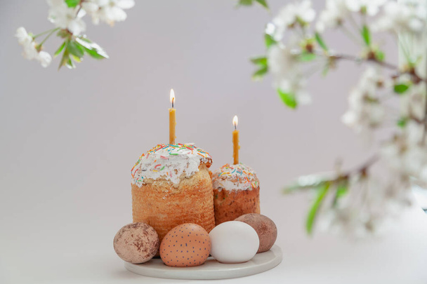 Easter still life. Two Easter cakes and eggs stand on a white background. Burning candles are inserted into the cakes. Top view of a tree branch with white flower - Фото, изображение