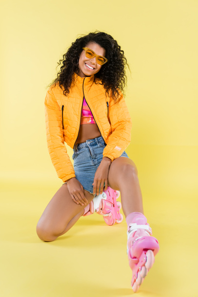 full length of smiling african american young woman in sunglasses and pink roller skates sitting on yellow - Foto, Bild