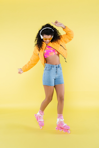 full length of african american young woman in headphones and pink roller skates on yellow - Foto, afbeelding