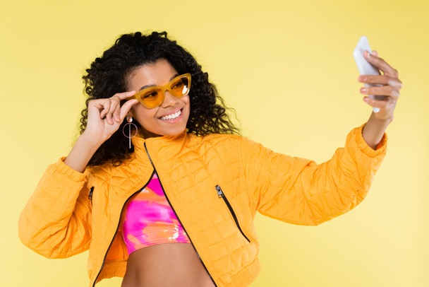 幸せなアフリカ系アメリカ人の若い女性がサングラスを調整しながら自撮りを黄色に隔離し - 写真・画像