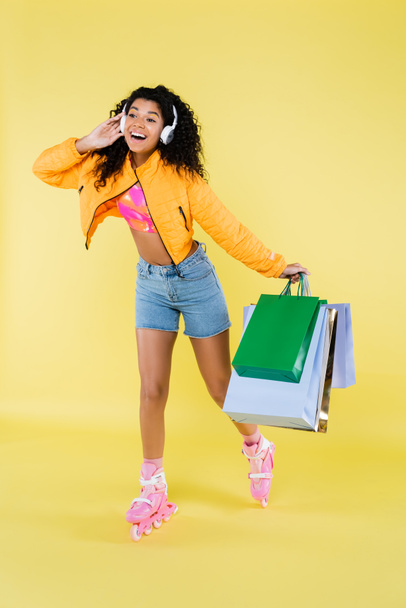 full length of amazed african american young woman in roller skates and headphones holding shopping bags on yellow - Fotoğraf, Görsel