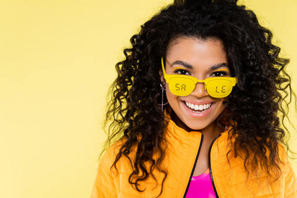 heureux afro-américain jeune femme en lunettes de soleil avec vente lettrage isolé sur jaune - Photo, image