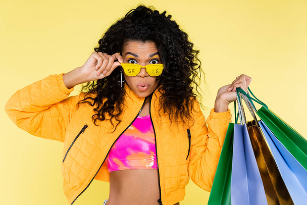 販売レタリング付きサングラスのアフリカ系アメリカ人の若い女性を驚かせた黄色に隔離されたショッピングバッグを保持 - 写真・画像
