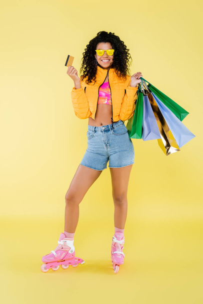 full length of happy african american woman in sunglasses and roller skates holding shopping bags and credit card on yellow - Foto, immagini