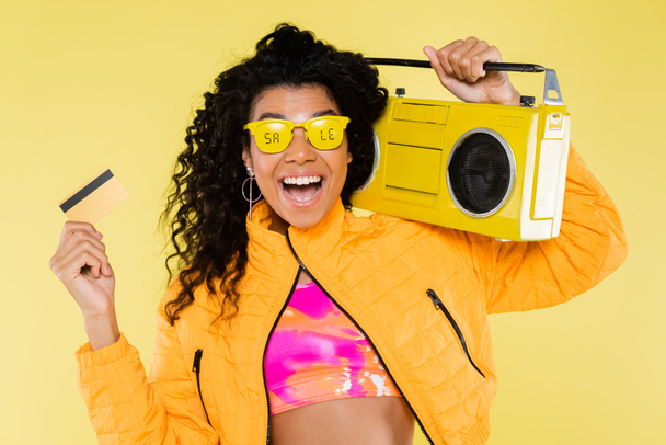 alegre Africano americano jovem mulher em óculos de sol com venda lettering segurando boombox e cartão de crédito isolado no amarelo - Foto, Imagem