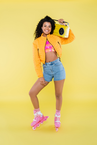 full length of happy african american woman on roller skates holding retro boombox on yellow - Foto, Imagem