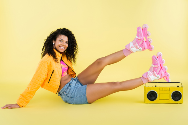 full length of positive african american woman on roller skates sitting near retro boombox on yellow - Photo, Image
